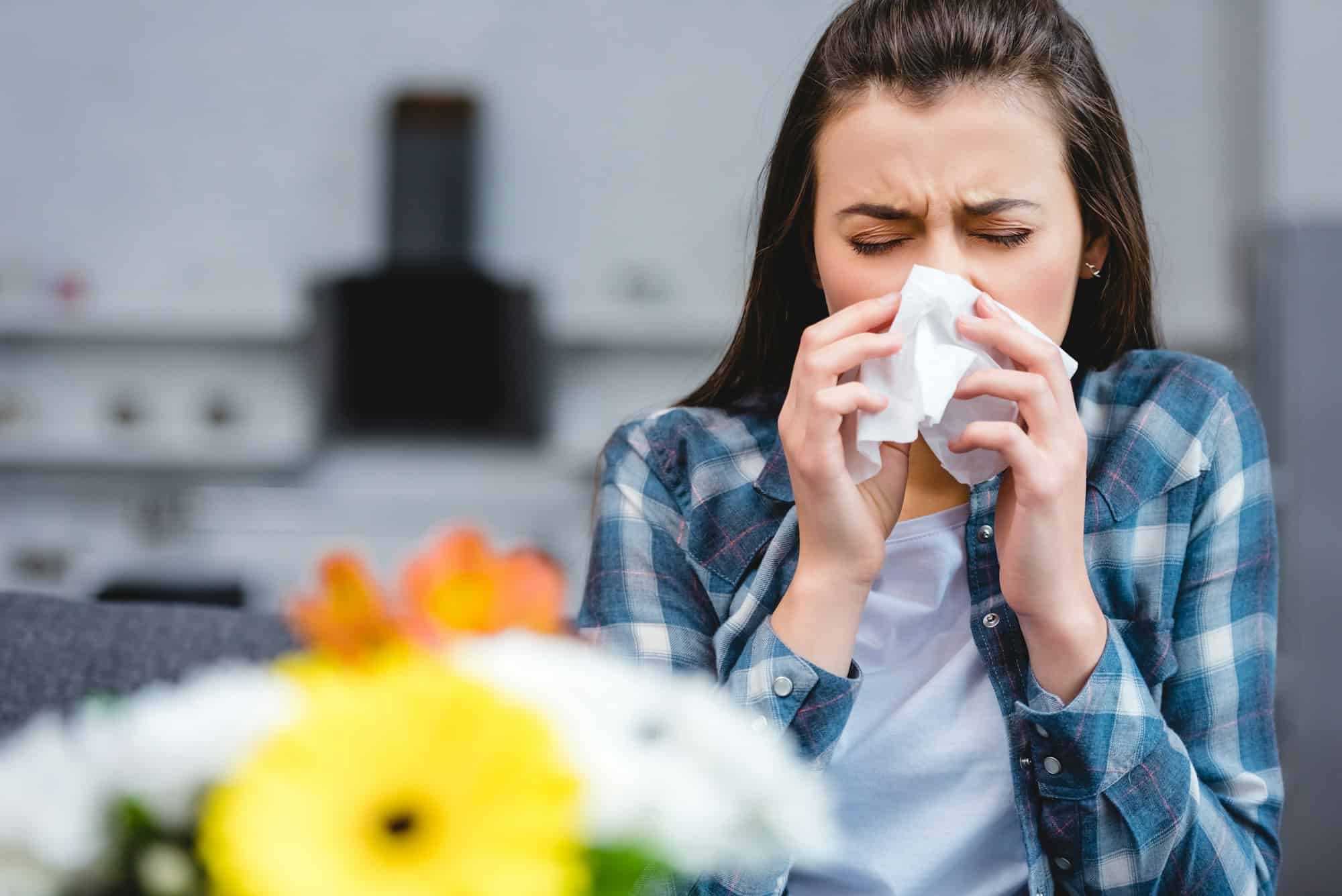 young woman with allergy holding facial tissue and sneezing, flowers on foreground